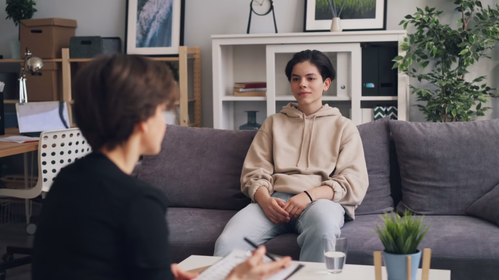 Teen client sitting on a couch during an in-person therapy counseling session with a mental health therapist in a calm office setting.