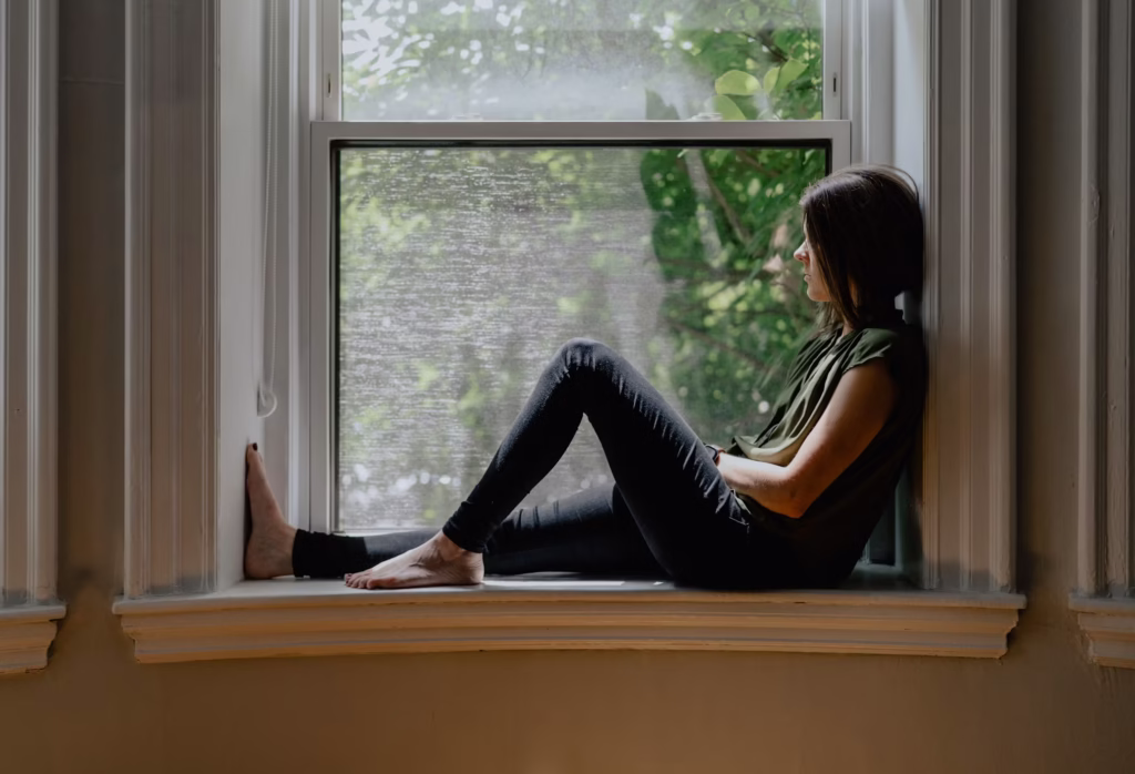 Person sitting thoughtfully in a window seat looking outside, surrounded by natural light and greenery, reflecting on emotions, healing, and personal growth.