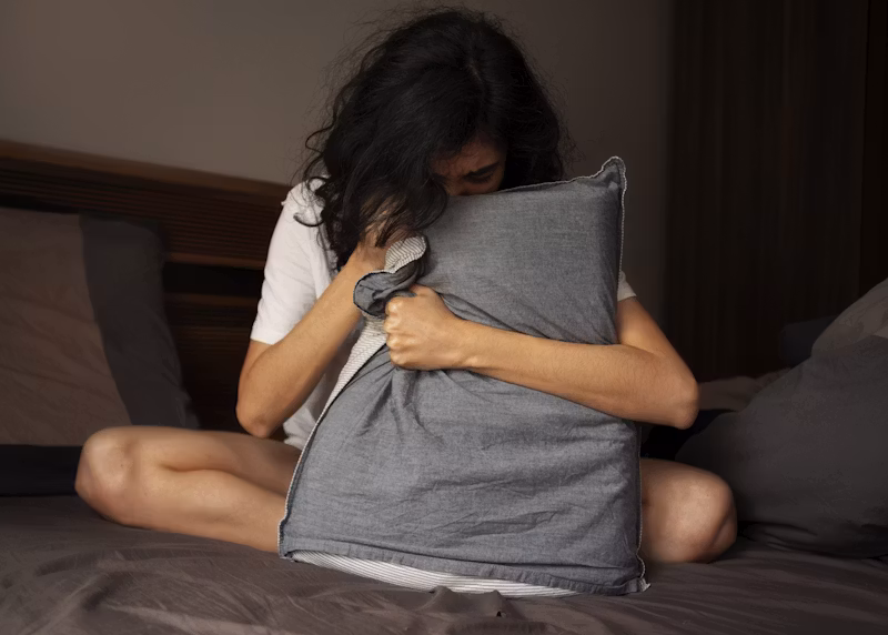 Person sitting on a bed hugging a pillow with their head down, representing anxiety, overwhelming worry, emotional distress, and the uncertainty that often accompanies anxiety disorders.